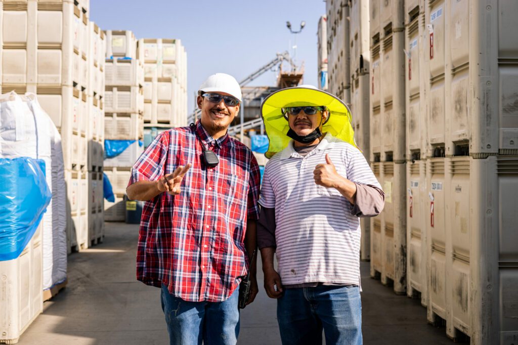 Two employees pose in front of stacked crates of almonds. Both are smiling and wearing tinted safety glasses and hard hats. The employee on the left is wearing a red plaid shirt and flashing a peace sign. His coworker is wearing a shade hat under his hard hat and has a striped polo over a brown long-sleeved shirt. He is giving a thumbs-up sign.