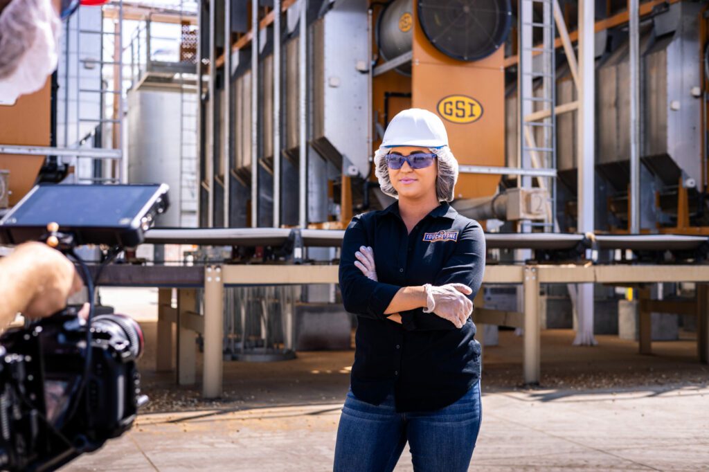 A Touchstone Pistachio employee stands in front of processing equipment. She is wearing a hairnet, tinted safety glasses, and a hard hat, along with a Touchstone Pistachio jacket and latex gloves. Her arms are crossed and she is smiling slightly.